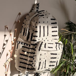Op de afbeelding: Een witte rugzak met een zwart geometrisch patroon hangt aan een muur. De rugzak heeft twee voorzakken en verstelbare bandjes. Het ontwerp heeft zwarte lijnen en stippen op een gewatteerde stof.