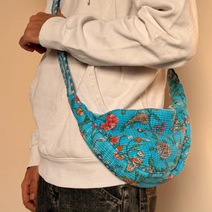 Könnte beinhalten: Türkisfarbene Umhängetasche mit floralem Muster in Rot, Orange und Grün. Die Tasche hat eine geschwungene Form und einen passenden Riemen. Der Hintergrund ist neutral.