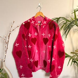 Puede incluir: Chaqueta de terciopelo rosa con cremallera, adornada con corazones rojos oscuros y cruces blancas. La chaqueta tiene cuello alto y está colgada de una percha de madera.