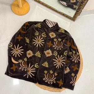 Puede incluir: Una chaqueta de terciopelo marr&oacute;n oscuro con cuello mao y mangas largas. La chaqueta est&aacute; decorada con motivos florales y abstractos bordados en tonos crema, verde claro y beige. El forro interior presenta un patr&oacute;n de rayas marrones y blancas.