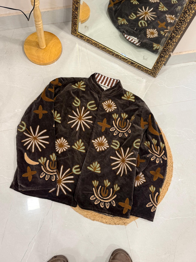 Puede incluir: Una chaqueta de terciopelo marr&oacute;n oscuro con cuello mao y mangas largas. La chaqueta est&aacute; decorada con motivos florales y abstractos bordados en tonos crema, verde claro y beige. El forro interior presenta un patr&oacute;n de rayas marrones y blancas.