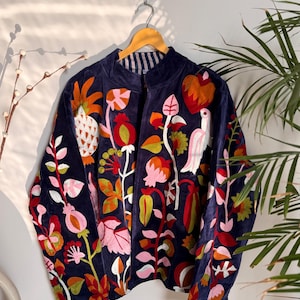 Puede incluir: Chaqueta azul marino con bordados florales y de pájaros coloridos. La chaqueta está colgada en una percha de madera contra una pared blanca. El bordado presenta detalles en naranja, rosa, rojo y verde, creando un diseño vibrante y artístico.