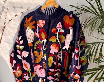 May include: A navy blue jacket with colorful floral and bird embroidery. The jacket is hanging on a wooden hanger against a white wall. The embroidery features orange, pink, red, and green details, creating a vibrant and artistic design.
