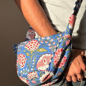 May include: A blue floral crossbody bag with a quilted design. The bag features a vibrant pattern of pink and red flowers with green leaves on a blue background. The strap is striped blue and white.