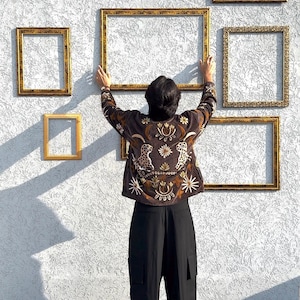 Puede incluir: Una chaqueta marr&oacute;n oscuro con motivos bordados de leopardos y flores en hilo dorado y blanco, combinada con pantalones negros. La chaqueta se sit&uacute;a frente a una pared decorada con m&uacute;ltiples marcos de fotos rectangulares dorados y vac&iacute;os.