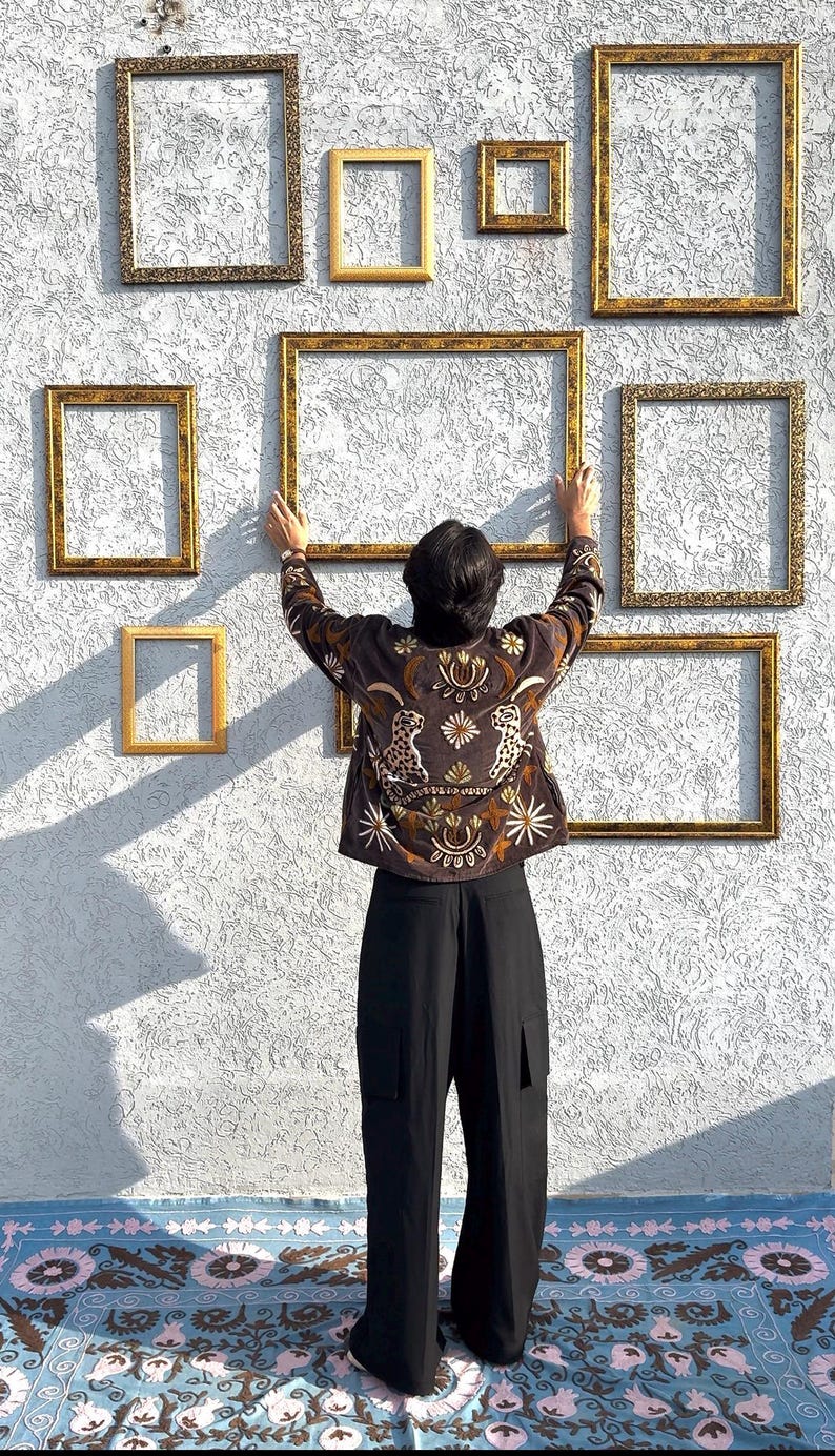 Puede incluir: Una chaqueta marr&oacute;n oscuro con motivos bordados de leopardos y flores en hilo dorado y blanco, combinada con pantalones negros. La chaqueta se sit&uacute;a frente a una pared decorada con m&uacute;ltiples marcos de fotos rectangulares dorados y vac&iacute;os.
