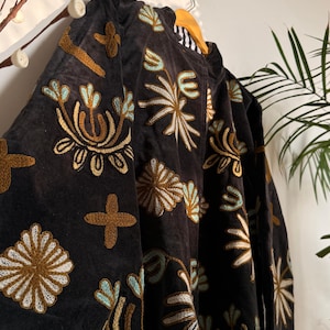 Puede incluir: Una chaqueta de color gris carb&oacute;n oscuro con bordados intrincados en tonos amarillo mostaza, azul claro y blanco. El dise&ntilde;o consiste en motivos florales repetitivos, formas de estrella y patrones de cruz sobre la tela. La chaqueta tiene cuello alto.