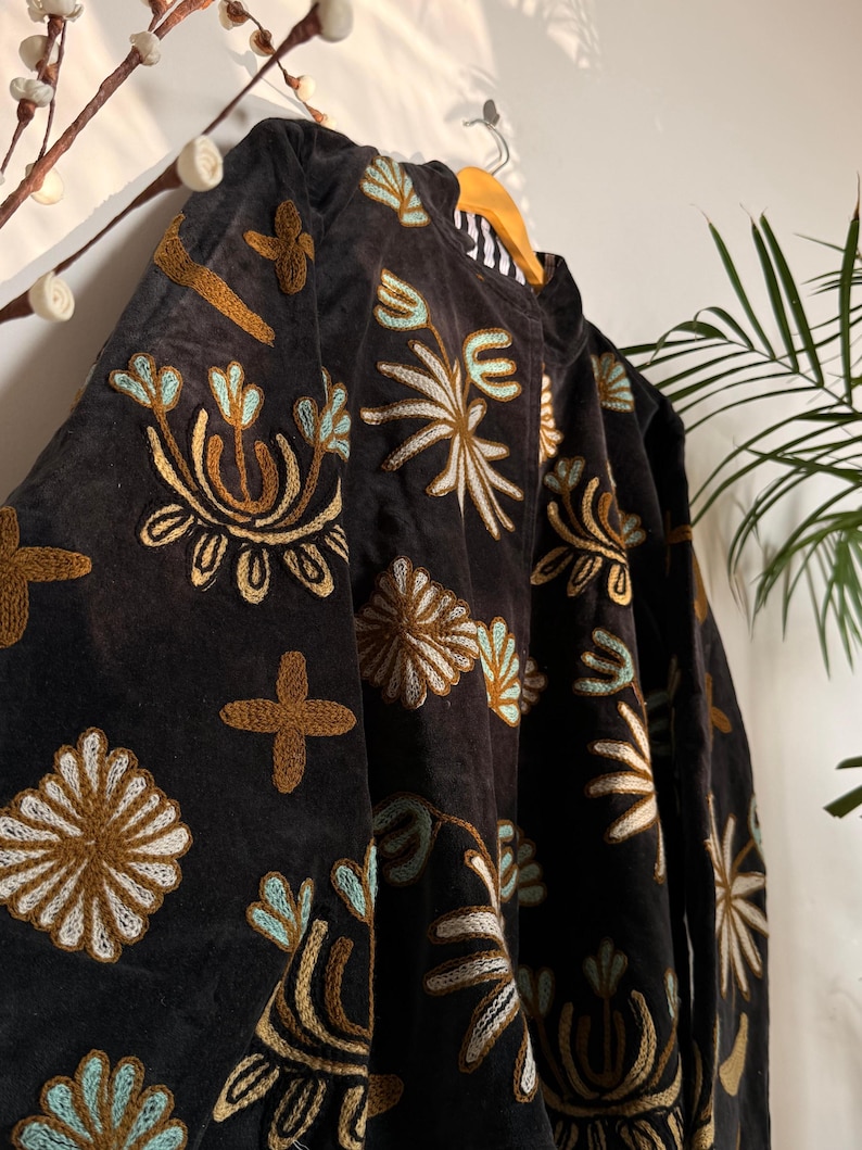 Puede incluir: Una chaqueta de color gris carb&oacute;n oscuro con bordados intrincados en tonos amarillo mostaza, azul claro y blanco. El dise&ntilde;o consiste en motivos florales repetitivos, formas de estrella y patrones de cruz sobre la tela. La chaqueta tiene cuello alto.