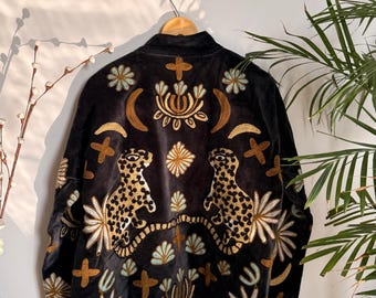 May include: Black velvet jacket with gold and cream embroidery. The jacket features two leopard figures and floral and cross designs. The jacket is hanging on a wooden hanger.