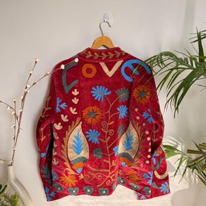 Puede incluir: Chaqueta de terciopelo rojo con bordados florales coloridos. La chaqueta tiene cuello alto y mangas largas. El bordado incluye diseños florales azules, naranjas y dorados, con la palabra "LOVE" bordada en la espalda. La chaqueta está colgada en una percha de madera.
