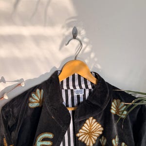 Puede incluir: Una chaqueta negra con patrones florales y geom&eacute;tricos bordados en tonos dorados y azul claro. La prenda tiene cuello, mangas largas y un forro de rayas verticales blancas y negras. Una etiqueta blanca interior indica 100% COTTON MADE IN INDIA FREE SIZE.