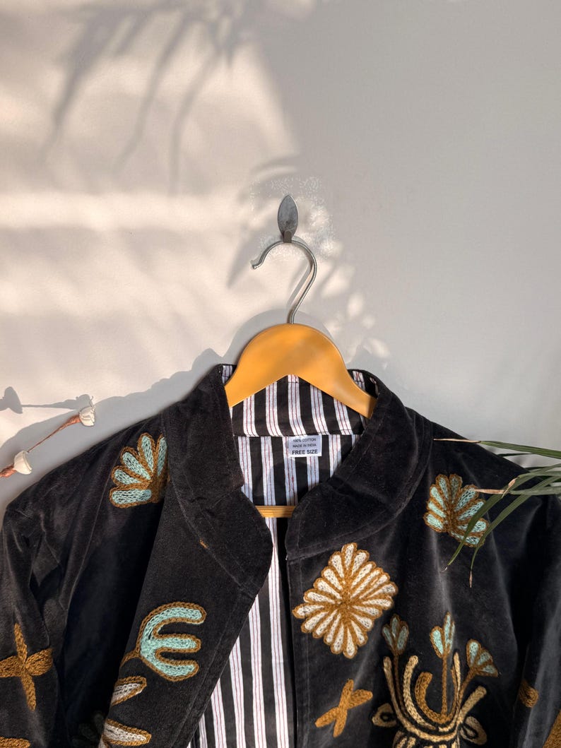Puede incluir: Una chaqueta negra con patrones florales y geom&eacute;tricos bordados en tonos dorados y azul claro. La prenda tiene cuello, mangas largas y un forro de rayas verticales blancas y negras. Una etiqueta blanca interior indica 100% COTTON MADE IN INDIA FREE SIZE.