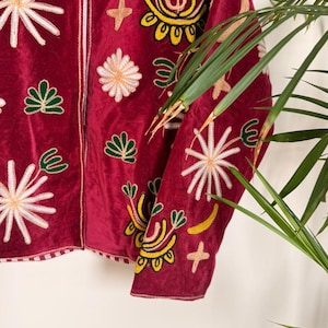 Puede incluir: Una chaqueta de terciopelo rojo oscuro con bordados intrincados en tonos amarillo, blanco y verde. El dise&ntilde;o incluye varios motivos florales y en forma de estrella repartidos por el frente y las mangas, con un borde a rayas verticales.