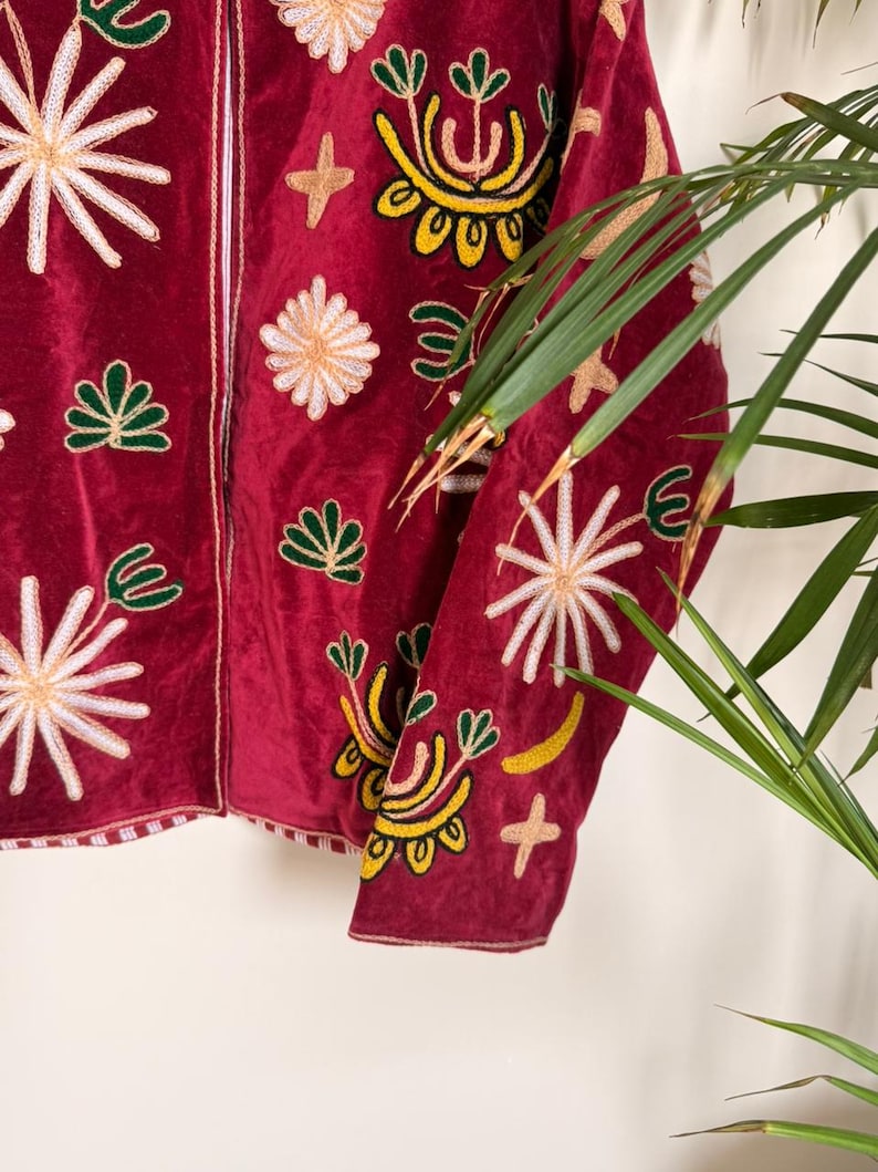 Puede incluir: Una chaqueta de terciopelo rojo oscuro con bordados intrincados en tonos amarillo, blanco y verde. El dise&ntilde;o incluye varios motivos florales y en forma de estrella repartidos por el frente y las mangas, con un borde a rayas verticales.