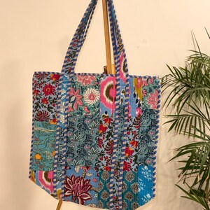 Op de afbeelding: Een patchwork tote bag met een bloemenpatroon in tinten blauw, roze en rood. De tas heeft twee handvatten met een gestreept ontwerp. De tas is gemaakt van gewatteerde stof en wordt tentoongesteld op een houten ezel.