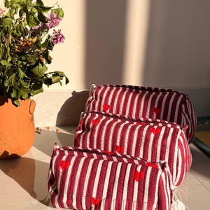 以下が含まれることがあります： 赤と白のストライプ模様のコスメポーチ3つ。赤いハートのアクセント付き。バッグは重ねられており、一番小さいバッグにはタッセルが付いています。背景には植物が入ったテラコッタの鉢があります。