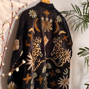 Puede incluir: Una chaqueta de color gris carb&oacute;n oscuro con intrincados bordados de dos leopardos, lunas crecientes y varios motivos florales y en forma de cruz en tonos dorados, azul claro y blanco. La prenda tiene un cuello alto y mangas largas.