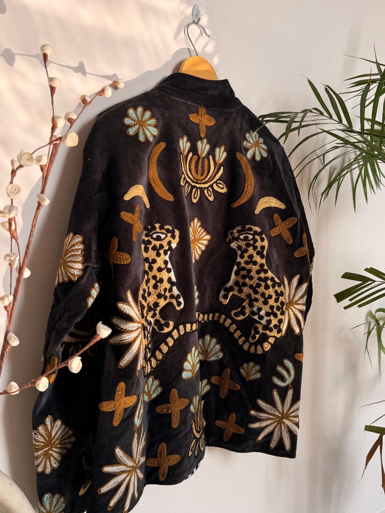 Puede incluir: Una chaqueta de color gris carb&oacute;n oscuro con intrincados bordados de dos leopardos, lunas crecientes y varios motivos florales y en forma de cruz en tonos dorados, azul claro y blanco. La prenda tiene un cuello alto y mangas largas.