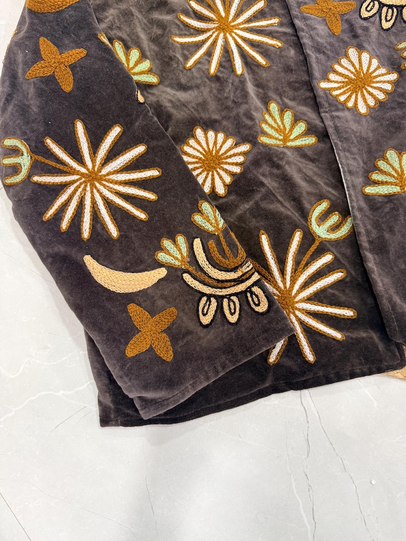 Puede incluir: Una chaqueta de terciopelo gris oscuro con bordados intrincados en tonos amarillo mostaza, blanco y verde menta claro. El dise&ntilde;o incluye varios motivos de estrellas, flores y formas de media luna sobre la tela.