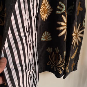 Puede incluir: Una chaqueta gris oscuro con intrincados bordados de flores y formas abstractas en tonos dorados, blancos y azul claro. El forro interior es de tela con rayas verticales blancas y negras, visible a lo largo del borde frontal abierto.
