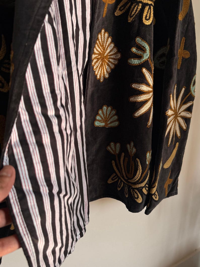 Puede incluir: Una chaqueta gris oscuro con intrincados bordados de flores y formas abstractas en tonos dorados, blancos y azul claro. El forro interior es de tela con rayas verticales blancas y negras, visible a lo largo del borde frontal abierto.