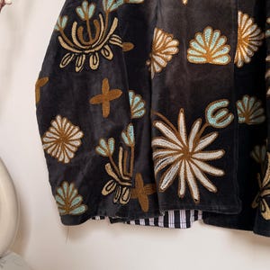 Puede incluir: Una chaqueta de terciopelo gris carb&oacute;n oscuro con intrincados bordados en tonos azul claro, crema y marr&oacute;n dorado. El bordado representa varias formas florales y abstractas sobre la tela. La chaqueta tiene un forro a rayas en blanco y negro.