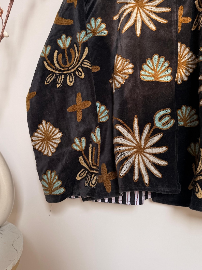 Puede incluir: Una chaqueta de terciopelo gris carb&oacute;n oscuro con intrincados bordados en tonos azul claro, crema y marr&oacute;n dorado. El bordado representa varias formas florales y abstractas sobre la tela. La chaqueta tiene un forro a rayas en blanco y negro.