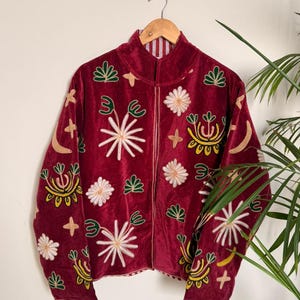Puede incluir: Una chaqueta de terciopelo rojo oscuro con mangas largas, cuello alto y cierre de cremallera frontal. La tela est&aacute; decorada con motivos bordados que incluyen estrellas blancas, formas florales verdes, lunas crecientes amarillas y cruces doradas.