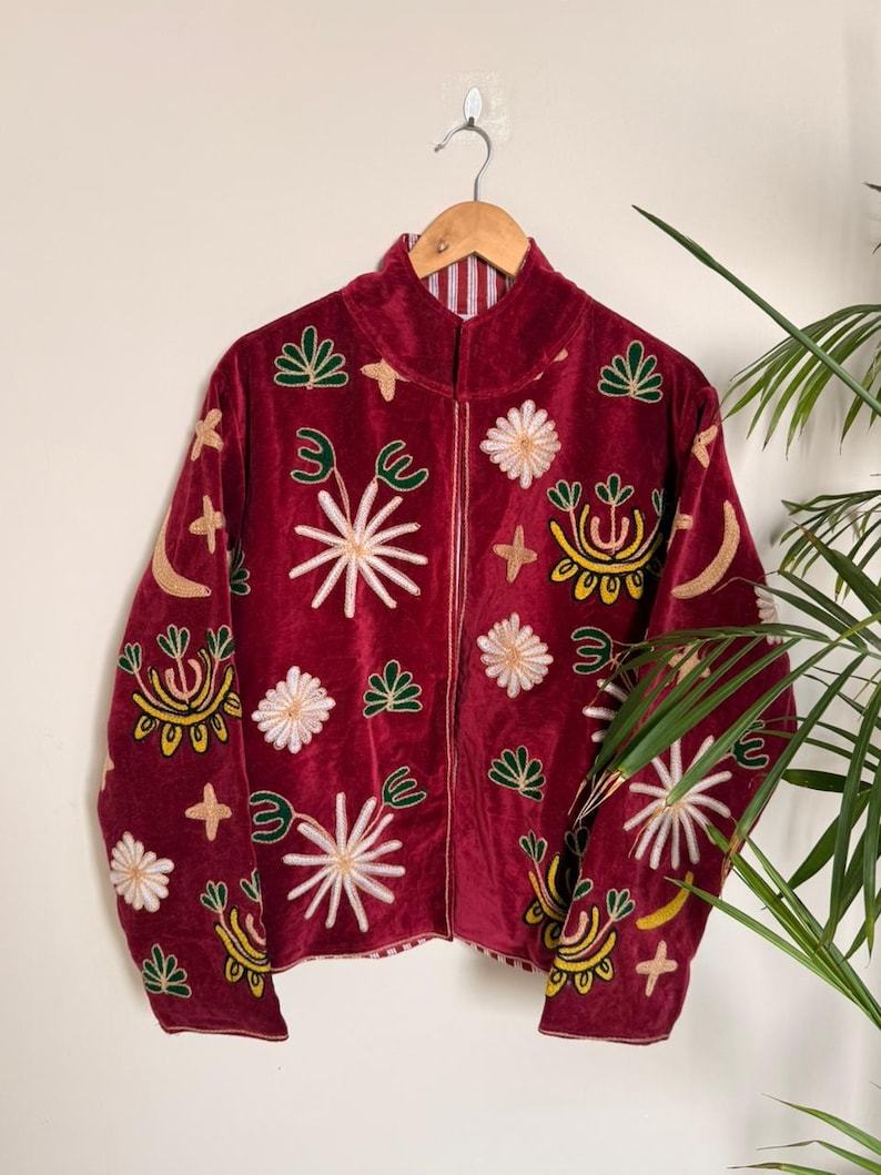 Puede incluir: Una chaqueta de terciopelo rojo oscuro con mangas largas, cuello alto y cierre de cremallera frontal. La tela est&aacute; decorada con motivos bordados que incluyen estrellas blancas, formas florales verdes, lunas crecientes amarillas y cruces doradas.