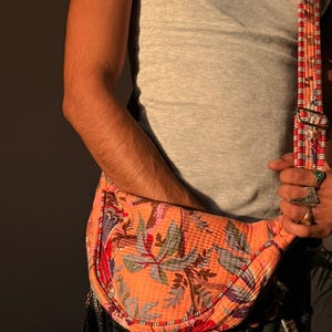 May include: A quilted orange fanny pack with a floral pattern in shades of pink, green, and blue. The bag has a matching patterned strap and a silver-toned buckle. The person is wearing a gray tank top and dark jeans.