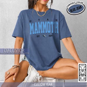 May include: A blue jean-coloured t-shirt featuring the text "UTAH MAMMOTH" in a bold, two-tone font. The shirt includes "EST. 2024" and a logo. The words "COMFORT COLOR" and "BLUE JEAN" are in a circular design. The bottom of the shirt says "CUSTOM YEAR".
