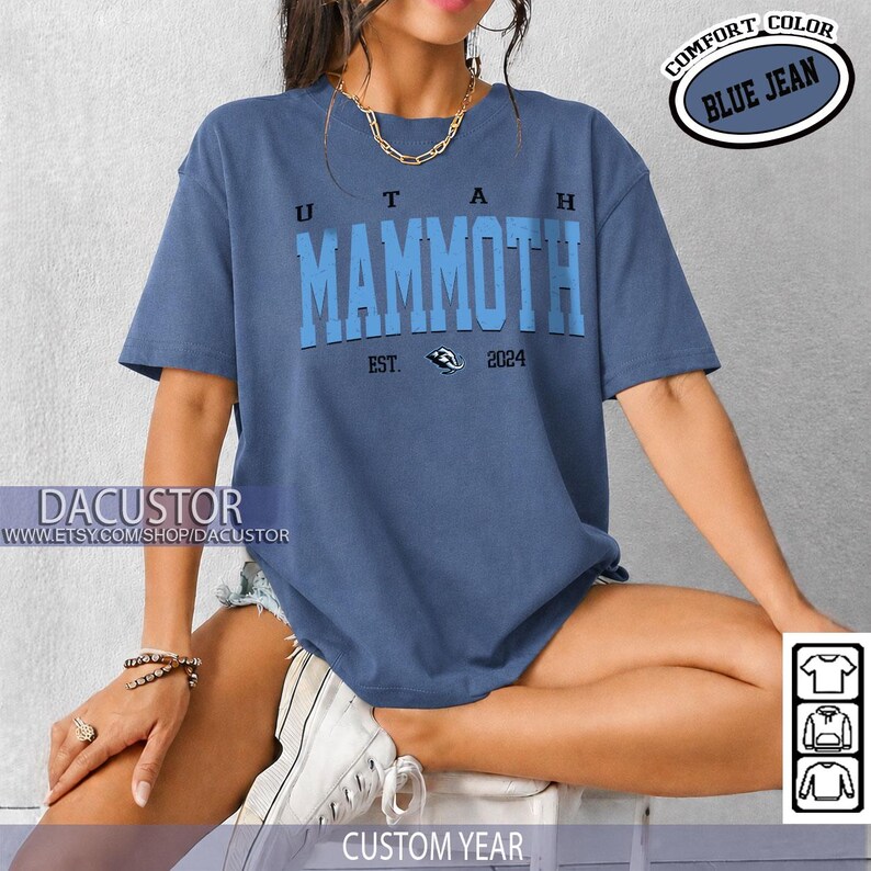 May include: A blue jean-coloured t-shirt featuring the text "UTAH MAMMOTH" in a bold, two-tone font. The shirt includes "EST. 2024" and a logo. The words "COMFORT COLOR" and "BLUE JEAN" are in a circular design. The bottom of the shirt says "CUSTOM YEAR".