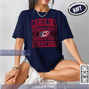 May include: Navy blue t-shirt with the text "CAROLINA HURRICANES" in red. The shirt features a logo with a red and black checkered pattern and the text "EST. 1997". The shirt also has the text "COMFORT, COLOR NAVY".