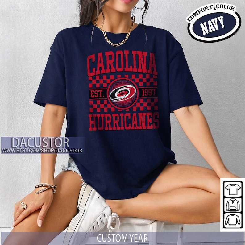 May include: Navy blue t-shirt with the text "CAROLINA HURRICANES" in red. The shirt features a logo with a red and black checkered pattern and the text "EST. 1997". The shirt also has the text "COMFORT, COLOR NAVY".