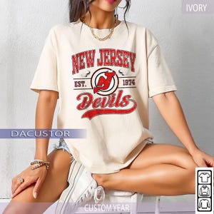 May include: Ivory-colored t-shirt featuring a distressed red and white graphic of the New Jersey Devils logo, with the text "NEW JERSEY" above and "Devils" below. The shirt includes the text "EST. 1974".