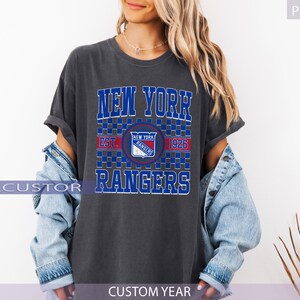 May include: Dark gray t-shirt featuring the text "NEW YORK RANGERS" in blue and white block letters. The shirt includes a team logo and the year "1926". The shirt is worn with a light blue denim jacket.