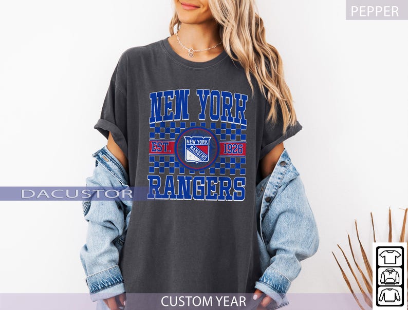 May include: Dark gray t-shirt featuring the text "NEW YORK RANGERS" in blue and white block letters. The shirt includes a team logo and the year "1926". The shirt is worn with a light blue denim jacket.