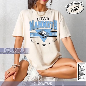 K&ouml;nnte beinhalten: Elfenbeinfarbenes T-Shirt mit dem Text "Utah Mammoth" in Blau, mit einer Mammut-Grafik in einem Dreieck. Das Shirt enth&auml;lt den Text "EST. 2025" und die Worte "Comfort Color Ivory". Der Etsy-Shop-Name "DACUSTOR" ist ebenfalls sichtbar.