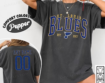 May include: A charcoal grey short-sleeve t-shirt featuring the text ST. LOUIS BLUES EST. 1967 in blue and gold lettering on the front. The back of the shirt displays the text ANY NAME above the number 00 in blue with gold outlines.