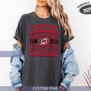May include: Dark gray t-shirt with the text "CAROLINA HURRICANES" in red, with a logo and the year "1997". The shirt is a Comfort Color Pepper brand.