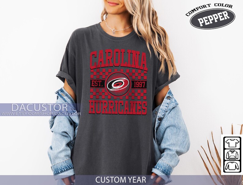 May include: Dark gray t-shirt with the text "CAROLINA HURRICANES" in red, with a logo and the year "1997". The shirt is a Comfort Color Pepper brand.