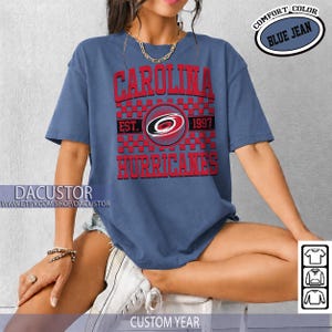 May include: Blue jean-colored t-shirt with the text "CAROLINA HURRICANES" in red. The shirt features the team's logo and the text "EST. 1997". The shirt has short sleeves and a crew neck. The words "COMFORT COLOR" and "BLUE JEAN" are also visible.