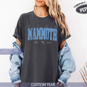 May include: A dark grey t-shirt featuring the text "UTAH MAMMOTH" in light blue, with "EST. 2024" below. The shirt is part of a clothing collection, and the words "COMFORT COLOR PEPPER" are printed on the top right.
