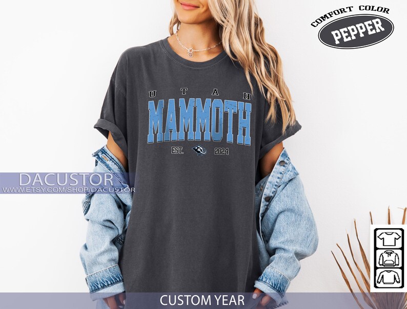 May include: A dark grey t-shirt featuring the text "UTAH MAMMOTH" in light blue, with "EST. 2024" below. The shirt is part of a clothing collection, and the words "COMFORT COLOR PEPPER" are printed on the top right.