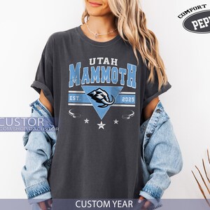 K&ouml;nnte beinhalten: Dunkelgraues T-Shirt mit dem Text "Utah Mammoth" in Hellblau und einer Mammut-Grafik in einem blauen Dreieck. Das Shirt enth&auml;lt das Jahr "2025" und die Worte "Comfort Color Pepper". Teil der "Custom Year"-Kollektion.