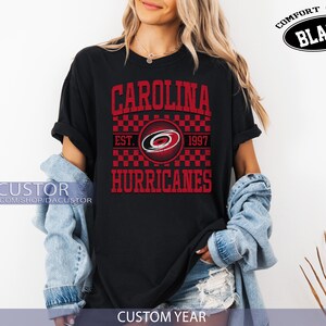 May include: Black t-shirt with a red and white graphic design. The design features the text "CAROLINA HURRICANES" with the team logo and "EST. 1997". The shirt is labeled "COMFORT COLOR BLACK" and "CUSTOM YEAR".