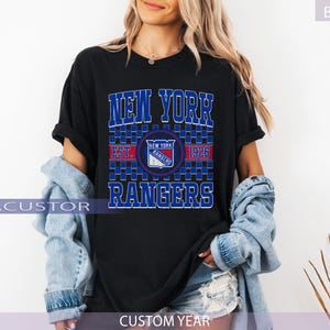May include: Black t-shirt featuring the text "NEW YORK RANGERS" in blue, with a team logo and "EST. 1926". The shirt has a classic design, perfect for fans of the hockey team. The shirt is a comfortable fit.