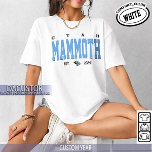 May include: White t-shirt with the text "UTAH MAMMOTH" in light blue block letters. The shirt also features the text "EST. 2024" and a Mammoth logo. The words "COMFORT, COLOR WHITE" are in a circle on the top right.