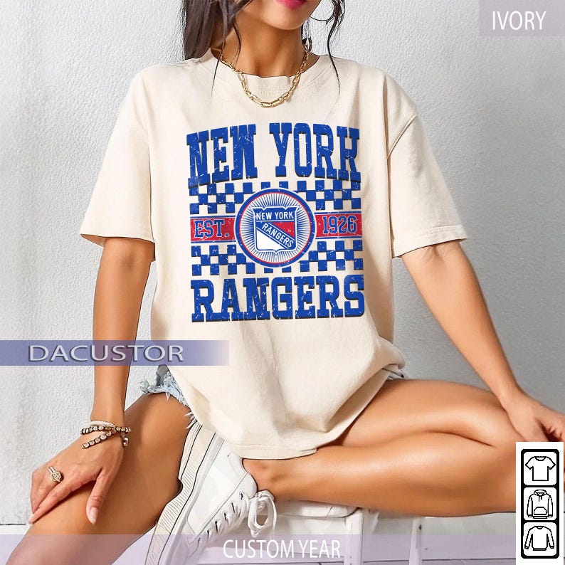 May include: Ivory-colored t-shirt featuring the text "NEW YORK RANGERS" in blue, with a checkered pattern. The shirt includes a circular logo with "NEW YORK RANGERS" and "EST. 1926". The word "DACUSTOR" is printed on the bottom left.