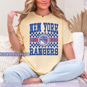 May include: A light yellow t-shirt featuring the text "NEW YORK RANGERS" in blue block letters. The shirt includes a circular logo with the team name and "EST. 1926". The shirt is a comfortable fit.